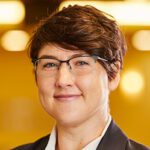 A woman with short hair and glasses, no earrings , a black blazer smiles at the camera in front of a brown and orange blurred background.